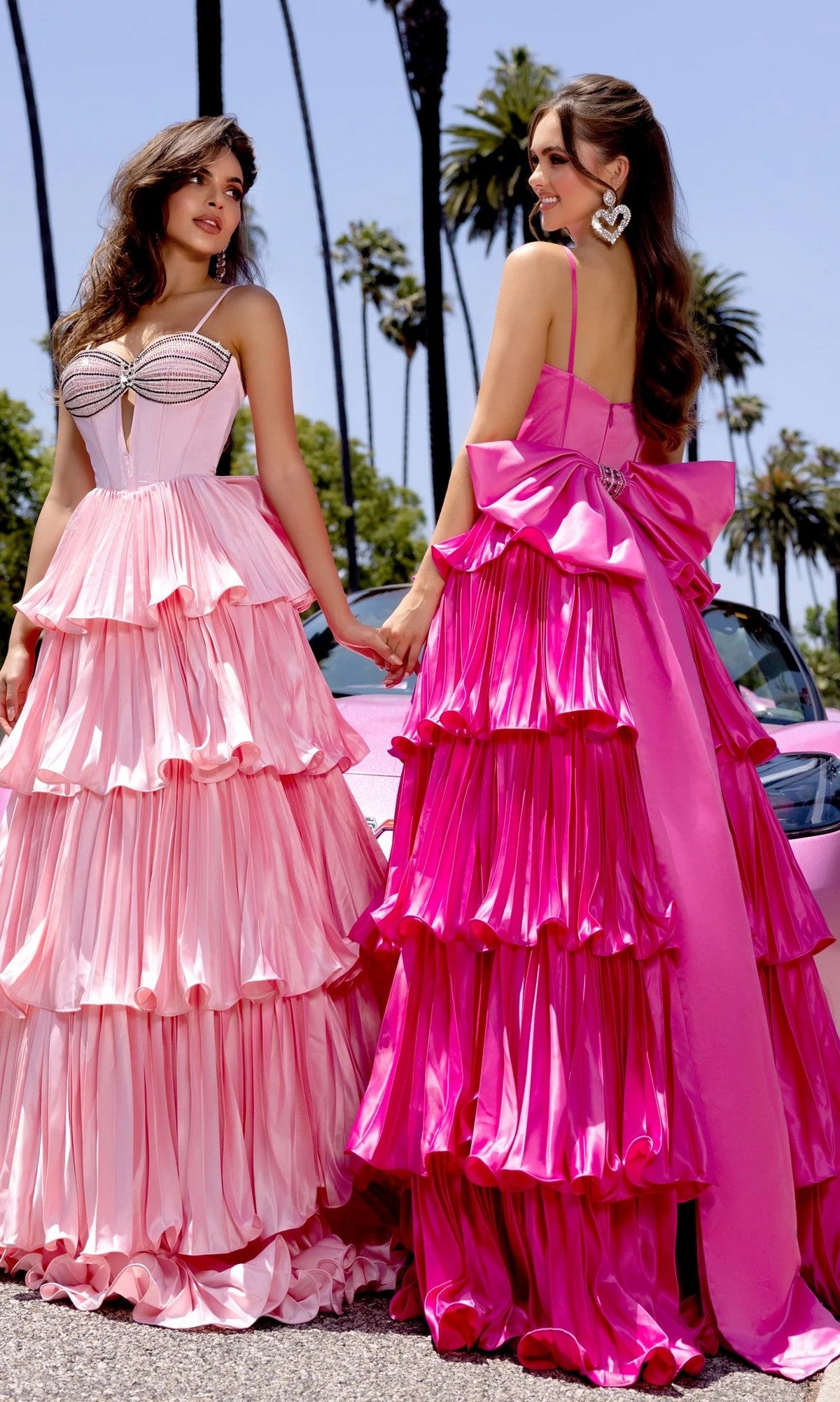 Two women wearing Portia and Scarlett PS25165 long formal dresses in hot pink stand outdoors holding hands, with palm trees and a pink car in the background.